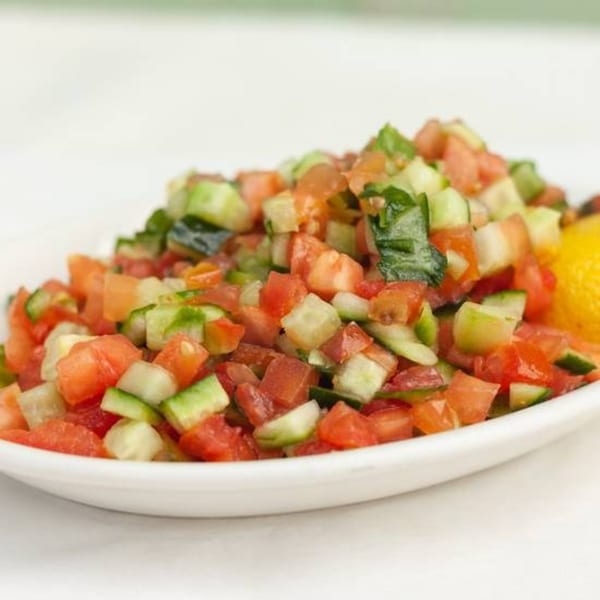 A white bowl filled with a fresh Israeli salad of diced tomatoes and cucumbers, with a lemon wedge.