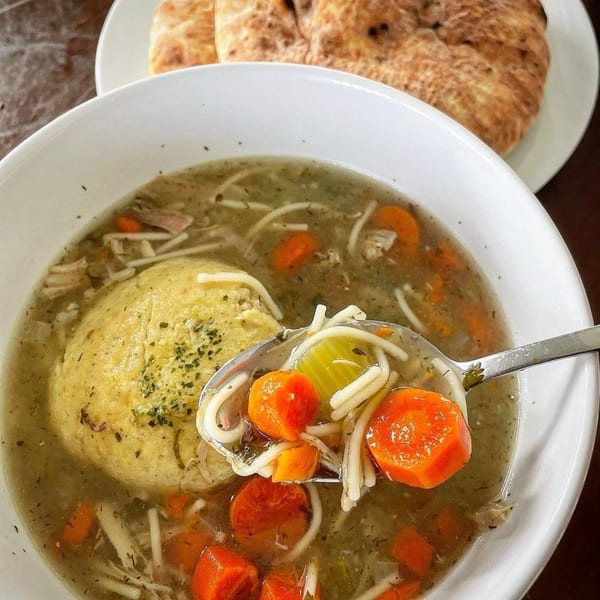 A bowl of matzo ball soup with carrots, noodles, and a large matzo ball, with pita bread in the background.