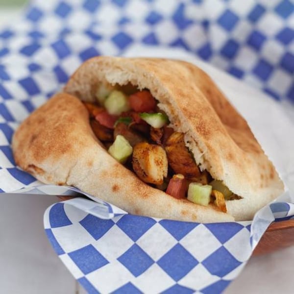 A pita bread sandwich filled with chicken, diced tomatoes, and cucumbers, served in blue and white checkered paper.